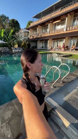 A woman in a black workout outfit stands by a swimming pool on a sunny day, holding a glass water bottle. She is in front of a multi-story building with several windows and outdoor seating areas. The pool has a tile bottom and is surrounded by lounge chairs and lush greenery, including trees and tropical plants. The woman looks at the camera and appears to be taking a selfie. The camera then pans to show a clearer view of the pool and the surrounding environment, highlighting the trees and landscape beyond the pool’s edge.