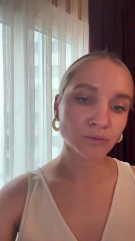 A close-up shot of a woman applying makeup is shown. She has blonde hair pulled back, light-colored eyes, and fair skin. She is wearing a light-colored, possibly off-white, sleeveless top. Prominent yellow hoop earrings are visible in both ears, and she has a small nose ring.

The background features a window covered by sheer, light-colored curtains, with a hint of an urban building visible outside. To the right of the frame, a dark red curtain is visible.

At the beginning of the video, she is looking slightly downwards. She then looks up, holding a dark, pen-like makeup applicator in her right hand and a small, white cylindrical object in her left. She brings the dark applicator to her right eyebrow and begins to apply makeup, focusing intently. After a few seconds of applying the product, she looks directly into the camera with a neutral expression. The lighting is bright, coming primarily from the window.