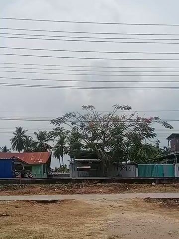 The video shows a landscape scene under a cloudy sky. Overhead, numerous power lines stretch across the frame. A medium-sized tree with a mix of green foliage and some reddish flowers is centrally positioned. Behind the tree, there are several structures, including a small, dilapidated house with a rusty-looking roof, a building with a white wall and some graffiti, and a teal-colored fence.

To the left of the tree, a few palm trees are visible. In the foreground, there is a dirt ground, and running parallel to the buildings is a set of railroad tracks.

A person rides a small motorcycle behind the tree, moving from left to right across the frame.