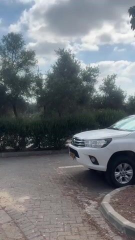 The video shows a white Toyota Hilux parked on the left side of a paved area, with trees and bushes in the background. The camera pans to the right, revealing a line of parked cars, including a silver sedan and a dark-colored sedan. Further to the right, there's a large palm tree and a no-entry signpost. The ground is covered with a mix of paving stones and dirt. The sky is visible with some clouds.