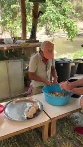 Two men are making balls of some kind of food. One man is sitting on a low bench, wearing a white shirt, a tan baseball cap, and a red mask around his neck. He is taking handfuls of the food mixture out of a large, blue plastic bowl and rolling it into balls. He places the balls on a large, silver metal tray. The second man is standing next to the seated man and is also taking handfuls of the food mixture and rolling it into balls. He is wearing a black hat and red shoes. The men are working on a wooden table that is sitting on a patterned rug. Behind them is a small bamboo structure with a metal tank and food containers. There is a river visible in the background. The scene appears to be outdoors under a covered area.
