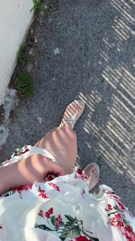 The video shows a first-person perspective of someone walking on a paved surface. The person is wearing a white floral dress and white sandals. Sunlight casts striped shadows on the ground. To the left, there is a light-colored wall and some sparse green vegetation growing at its base. The person's legs and feet are visible as they walk forward, and the camera angle is looking down. The overall impression is of a casual outdoor setting, possibly a sidewalk or path.