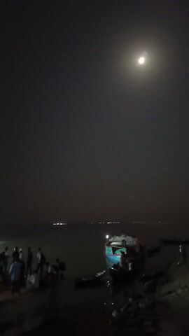 At night, a crowd of people gathers near a pier as a large, lit ferry approaches. A smaller boat is visible to the right. The moon is visible in the sky. The water is calm. People are standing, walking, and sitting. A small structure with a roof is on the pier. Lights are visible in the distance across the water.