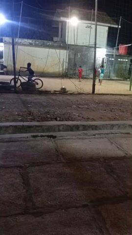 A group of children are playing volleyball on an outdoor court at night. The court is lit by a single overhead light. In the foreground, a person is riding a bicycle. The court appears to be made of dirt or gravel, and there is a net set up for the game. In the background, there are some buildings. The children are wearing casual clothing.