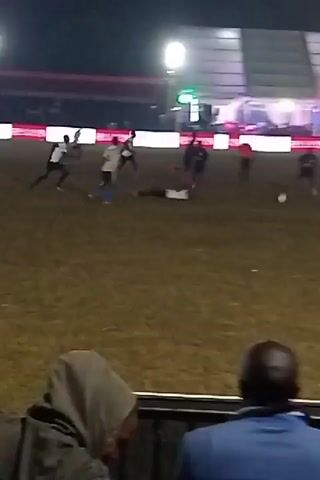 At night, a soccer game is taking place on a grass field. Several players in blue and white jerseys are running and kicking the ball. Behind the players is a large white tent with illuminated signs that read "CLEO MALALA" and "HOLDIN." There are several spectators watching the game, one of whom is wearing a blue jacket. The lighting is bright, casting shadows on the field.