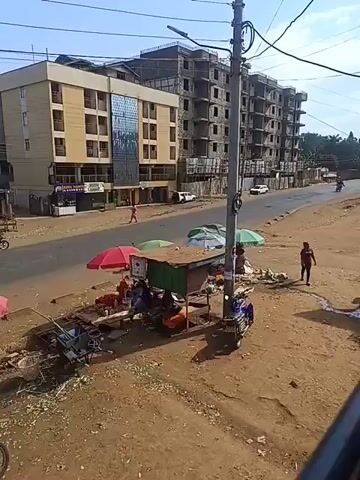 The video shows a street scene in an urban area, likely in Africa. A two-lane road runs through the center of the frame, with vehicles like motorcycles and a large white truck passing by. On the right side of the road, there's a dirt area with several small market stalls sheltered by colorful umbrellas. People are seen sitting and standing around the stalls, possibly selling goods. In the background, there are buildings including a tan-colored multi-story building and a building under construction. Utility poles with electrical wires are visible, adding to the urban landscape. The sky is blue and mostly clear. The overall scene suggests a busy, everyday environment in a developing urban area.