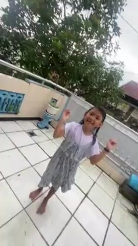 A young girl is standing barefoot on a white tile patio, looking directly at the camera and speaking. She has dark hair in braids and is wearing a white t-shirt under a sleeveless, patterned gray dress. She is smiling and gesturing with her hands as she talks. Behind her, there is a low concrete wall topped with a metal railing. A green tree is visible behind the wall. In the background, there is a glimpse of a neighboring house with a red roof. The weather appears cloudy.