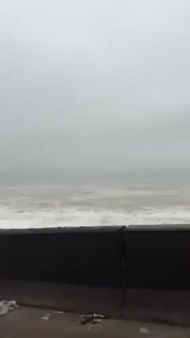 The scene captures a view of a stormy ocean on an overcast day. Waves crash against a stone barrier that separates the sea from a roadside. A car passes along the road in the distance. A solitary utility pole is present in the foreground, partially obscuring the view of the ocean. The overall atmosphere is bleak and the weather appears to be rough.