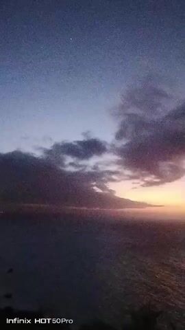 The video shows a time-lapse of a sunset over the ocean. The sky is a deep blue fading to pink and orange at the horizon. Clouds are present in the sky and are illuminated by the setting sun. The ocean is dark with ripples reflecting the light from the sky. There are some silhouetted trees in the foreground. A watermark at the bottom indicates the video was recorded on an Infinix HOT 50 Pro.