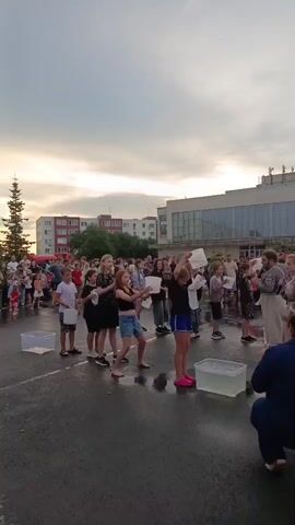 A group of children are participating in what appears to be an ice bucket challenge or a similar activity. The children are standing in a line outdoors in what looks like a paved area, possibly a schoolyard or town square. They are holding buckets or containers of water above their heads, and some have already poured the water over themselves. There are other people, presumably adults and a larger audience, watching the event. In the background, there are buildings and trees. The sky is overcast, suggesting it might be late afternoon or evening, and the ground is wet, indicating recent rain or the activity itself. The children are dressed in casual summer clothing. Some of the adults are filming the event with their phones.