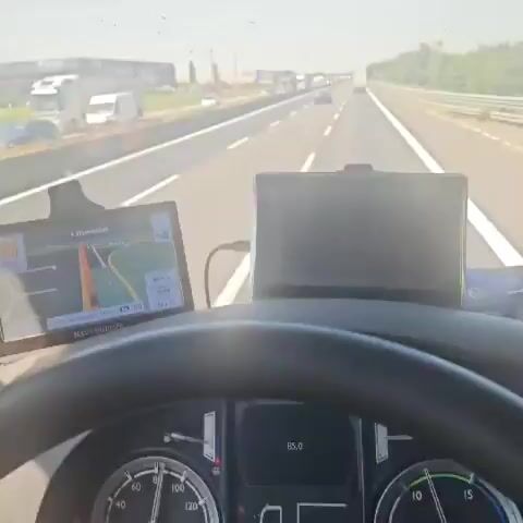 The video shows a view from the driver's seat of a vehicle moving along a highway on a bright, sunny day. The highway has multiple lanes and is filled with cars and trucks moving in the same direction. On the side of the highway, there is a large building with the word "Ferrari" visible, and another building with the words "PALFINGER ITALIA". The camera pans slightly to the left, showing more of the scenery along the highway, including trees and grassy areas. The vehicle appears to be moving at a steady pace. Two screens are visible in the lower portion of the shot, likely displaying navigation or vehicle information.
