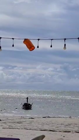 A string of lights, some with bulbs and one with an orange cylindrical object, hangs across the top of the frame. Below the string of lights, a calm, gray sea stretches to the horizon under a cloudy sky. A long, narrow boat with a canopy is anchored in the shallow water near a sandy beach. The boat appears to be empty or has a person inside who is not clearly visible. Another small red kayak is partially visible in the water to the right. The beach itself is sandy and appears to be mostly empty, with some debris scattered near the waterline. The overall atmosphere is peaceful and somewhat overcast.