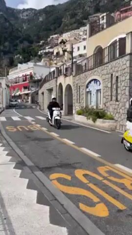 The video shows a street scene in a coastal town, possibly in Italy. A person wearing a helmet and dark clothing is riding a white scooter on the road. The road is marked with a yellow "BUS" lane. Buildings are terraced into the hillside, with a mix of architectural styles and colors, including white, red, and stone. Some buildings have arched features. The sky is partly cloudy and a mountain is visible in the background. The camera is stationary, capturing the scooter moving away.