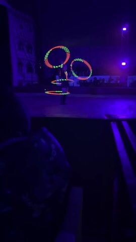 The video shows a person performing a hula hoop act under blacklights in a circus ring. The person has a hula hoop around their waist, and is spinning two more hoops simultaneously around their arms. The hoops glow orange and green. The circus ring is surrounded by tiered seating in front of a large banner. A few patrons are visible in the stands. The video is recorded from the lower bleachers, with a person wearing a dark blue patterned shirt visible in the foreground.