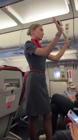 Here is a description of the video:
A flight attendant stands in the aisle of a plane, giving safety instructions with a mock demonstration of a seatbelt. She is wearing a gray uniform dress with a red scarf and belt. Her blonde hair is pulled back in a bun. She's holding a part of a seatbelt in both hands above her head. Passengers are seated and some are looking at their phones. The interior of the plane is visible with overhead compartments and emergency exit signs.
