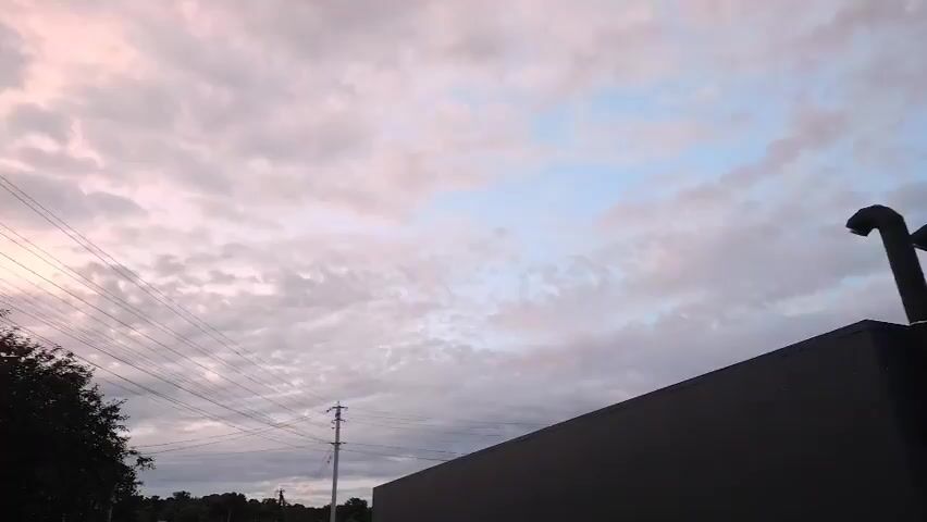 A low angle shot reveals a cloudy sky with a mix of white and pinkish clouds. In the foreground, the dark, angular roofline of a building is visible, adorned with three black exhaust pipes that curve downwards at the top. To the left, power lines stretch across the frame, supported by a utility pole. To the right, a section of a red, corrugated roof with a chimney protrudes into the frame. Trees are visible in the distance, silhouetted against the sky. The scene appears to be outdoors, likely during twilight.
