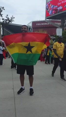 A man is standing on a sidewalk holding a Ghanaian flag. He is wearing a white t-shirt and black shorts. Behind him, there are people and a large screen that says "FIFA WORLD CUP 2026". The weather is overcast.