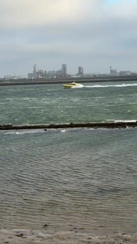 A yellow boat speeds across choppy green water, leaving a white wake. In the distance, a large industrial complex with several tall structures lines the horizon under a cloudy sky. The foreground shows a calm, shallow body of water with a sandy bottom, separated by a low barrier of rocks or debris from the rougher water beyond. The scene is filmed from a low vantage point near the shore.