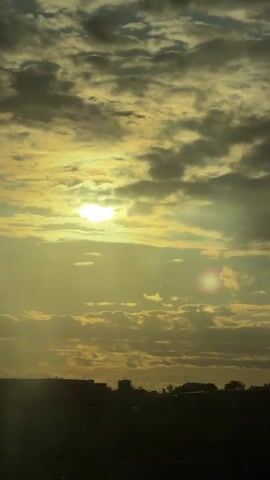 The video shows a sunset with a bright sun peeking through clouds. The sun is a bright white orb with a slightly yellow halo. The clouds are a mix of dark grey and light grey, with some yellow and orange hues. The bottom of the frame shows a silhouetted landscape with buildings and trees. The overall mood is calm and peaceful.
