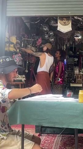 The video shows a person wearing a floral shirt and a bucket hat sitting at a table, occupied in drawing on paper. The table is covered with a green cloth and positioned on a patterned carpet. In the background, inside what appears to be a shop, a man wearing a white tank top and brown skirt is assisting a woman in a sequined top and patterned skirt. The shop interior is decorated with various items, including mannequins, jewelry, disco balls, and other decorative objects. The man in the tank top is handling some jewelry, while the woman smiles and interacts with him. The shop appears to be located beneath a partially open garage door, letting in natural light.