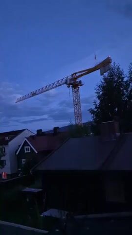 The video shows an exterior view of a construction crane at dusk. The sky is a deep blue with some clouds. The crane is yellow and white and stands tall against the sky. In the foreground, there are the rooftops of several houses, some with visible chimneys. Trees are also visible, some in the foreground and some behind the crane. The overall scene suggests a quiet, possibly suburban environment at the end of the day, with the presence of ongoing construction indicated by the crane. There are no people or animals visible in the footage. The lighting indicates it is either early evening or late evening.