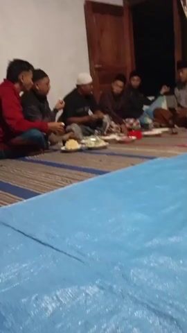 A group of men are sitting on the floor in what appears to be a room. They are gathered around several plates of food. Some of the men are wearing traditional clothing, including caps and sarongs. The room is simply furnished, with a blue tarp covering the floor in the foreground. The lighting suggests it is evening or nighttime. Some of the men are engaged in conversation, while others are eating. The overall atmosphere appears to be social and communal.