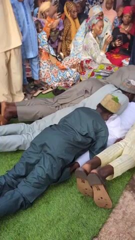 Four men are prostrating face down on a green artificial turf mat and a brown patterned mat. The men are dressed in traditional kaftans: one in dark green, one in white, one in light yellow, and one in black, each wearing a cap. In the background, a group of women are seated on the floor, observing the scene; they are dressed in various colorful traditional garments and head coverings. A woman in a black abaya and head covering stands behind the prostrating men, holding a microphone and appearing to speak. Another woman in a white lace-embroidered long-sleeved dress stands to the right of the woman with the microphone. The setting is decorated with white and pink draped fabric, suggesting an indoor or covered outdoor event.