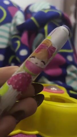 Here's a description of the video:

A hand with dark nails holds a pencil-shaped eraser with a cartoon design. The eraser has a purple cap. The background is blurred, showing a colorful patterned blanket and a yellow plastic toy with an opening on top. The video focuses on the eraser.