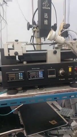 Here's a detailed description of the video, focusing on essential elements:

**Scene:** The video shows a stationary shot of a Chinatafix BGA rework station, likely in a workshop or electronics repair environment. 

**Objects:**
*   **BGA Rework Station:** The central focus is the machine itself. It has a black and silver color scheme, various control panels with digital displays, knobs, and buttons. Prominent labeling includes "Top Heater," "Bottom Heater," "IR," "Air Regulator," "Cool Fan," "Start," and "Stop." There is a heating head with a light.
*   **Workbench/Shelf:** The machine sits on a metal workbench or shelving unit.
*   **Electronic Components/Debris:** The area surrounding the machine is cluttered with various electronic components. A laptop screen, possibly disassembled, is visible.

**Environment:**
*   The setting looks like a workshop or repair area. The lighting is somewhat uneven, with a visible light source illuminating the machine.
*   The surrounding area is cluttered, suggesting a working environment focused on electronics repair.

**Action:**
*   The video does not capture anyone performing actions.
*   The machine is idle, with the display and potentially the fan running.

**Interactions:** There are no interactions with people in this video.

In summary, the video appears to be a demonstration or documentation of a BGA rework station within a repair or workshop setting.