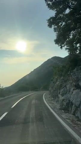 The video shows a point-of-view shot from a moving vehicle traveling along a paved road that curves to the right. The road has a white line on the left and right edges. To the right of the road, there is a steep, rocky embankment with some trees and vegetation growing on it. To the left of the road, there is a sloping hillside covered in trees and vegetation. The sun is visible in the upper left corner of the frame, partially obscured by clouds, casting a bright light on the scene. The sky is a clear blue with some scattered clouds. The overall impression is of a scenic drive through a mountainous, coastal area.