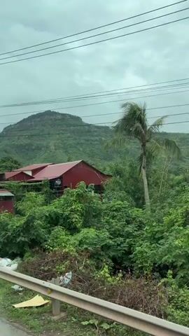 The video shows a series of roadside scenes from a moving vehicle. The first scene features a red-roofed building surrounded by lush greenery and a cloudy sky. The second scene reveals a sign with Vietnamese text, piles of bags, and more vegetation. The third scene displays a white house with a covered porch, a tree, and a sign with Vietnamese text. The fourth scene shows a small building with a gray metal gate and a cart, followed by a large tree and a motorcycle parked in front of a house. The overall environment appears to be rural or semi-rural, with a mix of residential and commercial establishments.