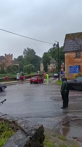 The video shows a flooded street in front of a building. It is raining heavily, and the road is covered in water. Several cars are visible, including a red car driving through the water and a grey car parked on the side of the road. Two people in raincoats are standing on the side of the road, watching the cars. In the background, there is a castle-like building and trees. A signpost indicates directions to various towns. The overall scene depicts a difficult driving situation due to the severe weather.