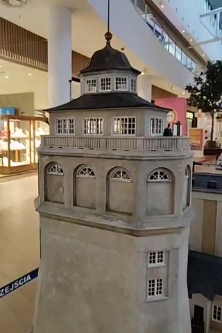 A close-up, panning shot showcases a detailed miniature model of a lighthouse situated inside a shopping mall. The lighthouse model is constructed with realistic textures and features multiple levels, windows, and a decorative railing. The top of the lighthouse is capped with a dark, multi-tiered roof. In the background, the bright lights and displays of various shops are visible, including a store with the "YES" logo. Shoppers can be seen in the distance, browsing or walking through the mall. The model includes a smaller, adjacent building with a dark roof and windows. The camera pans to reveal more of the mall environment, including signage and other retail displays.