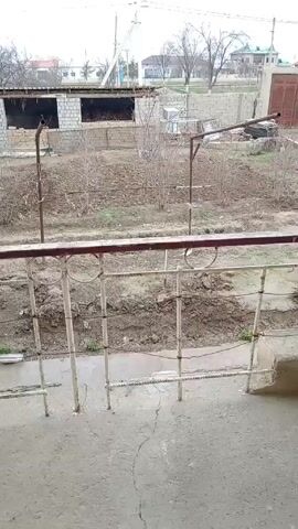 The video shows a view from a balcony overlooking a yard. The yard contains a partly constructed building, garden beds with dormant plants, and various objects scattered around. A metal fence runs along the edge of the balcony. Beyond the yard, there are houses, trees, and utility poles. The sky is overcast, suggesting a dull, grey day.