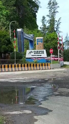 The video shows a wide shot of the entrance to a facility named "SOLUSI BANGUN ANDALAS Pabrik Lhoknga". In the foreground, there is a wet asphalt road with several puddles reflecting the sky and surrounding environment. A concrete barrier with yellow and black stripes separates the road from the facility entrance. The entrance features a large sign with the company name and logo, which appears to be a stylized letter "A". Behind the sign, there is a large sack with a logo that reads "ANDALAS". To the left and right of the sign are poles with Indonesian flags. A blue, U-shaped structure is visible behind the sign to the left, and a red stop sign is positioned to the right. The background consists of lush green trees and a rocky cliff face on the left, with taller trees on the right. The sky is overcast. The video appears to be a static recording of the entrance.