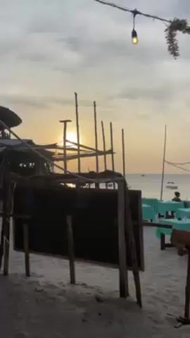 The video shows an outdoor restaurant on a sandy beach. The sun is setting over the water, creating a warm glow. There are several wooden tables covered with turquoise tablecloths. People are sitting at some of the tables, dining. There are decorations strung above the tables, including lights and decorative items. Boats are visible in the water in the background. A chalkboard stands near the entrance of the restaurant. The video pans slightly from left to right, revealing more of the restaurant and the surrounding beach.
