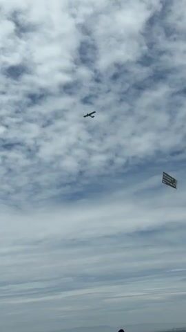 A small airplane flies across a blue sky filled with scattered white clouds. Attached to the airplane by a long rope is a rectangular banner. The banner is dark and appears to have text printed on it, but the text is not legible at this distance. In the lower part of the frame, a person's head is visible, and they are looking up at the sky.