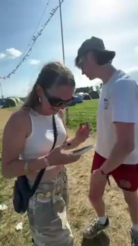 Here is a description of the video:

The video shows several people at what appears to be an outdoor event or festival. The scene features a large field filled with tents under a blue sky with scattered clouds.

The video first focuses on a woman with long hair wearing sunglasses, a white tank top, and patterned pants. She is looking at her phone. Then the video shows a man in a white t-shirt and red shorts holding a small container. A man in a hat and tank top is also visible, and briefly interacts with the woman. Overhead, there are lines of colorful flags. The ground is a mix of dirt and grass.