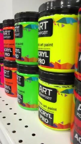 The video displays shelves stocked with jars of "ART KOMPOZIT ACRYL PRO" acrylic art paint. The jars are stacked in two rows, with various vibrant colors visible, including neon pink, orange, lime green, yellow, blue, and black. Each jar has a black lid and a label with the brand name, "ART KOMPOZIT," the product type "acrylic art paint," and "ACRYL PRO" in large letters. A colorful geometric pattern adorns the lower part of the labels. The setting appears to be a retail store or art supply shop. The lighting suggests an indoor environment, likely during the day. The camera pans across the shelves, offering a close-up view of the colorful paint jars, highlighting their uniformity and variety. There are no people visible in the frame, and the focus is solely on the products displayed. The overall impression is one of a well-organized display of art supplies, emphasizing the range of colors available.