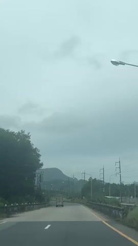 This media file captures a view from a vehicle traveling on a road in Tambon Takdat, Thailand. The scene is dominated by a gray, overcast sky, suggesting cloudy or possibly rainy weather. Lush green trees line the left side of the road, with some power lines and poles interspersed among them. On the right side of the road, more power lines and poles are visible, along with vegetation. A distinctive mountain range with rounded peaks is visible in the distance, partially obscured by the clouds.

The road itself is paved and appears to be a two-lane highway. White dashed lines mark the lanes, and a solid yellow line delineates the right edge of the road. A single white truck is seen driving ahead in the same direction as the viewer. There are also a few streetlights along the roadside, and some yellow directional signs indicating curves in the road. The overall impression is one of a somewhat rural or semi-urban drive on a gloomy day. The presence of the power lines and the type of vegetation might suggest a tropical or subtropical climate. There is no discernible text visible in the media. The activity is the act of traveling by road, with the camera capturing the passing scenery.