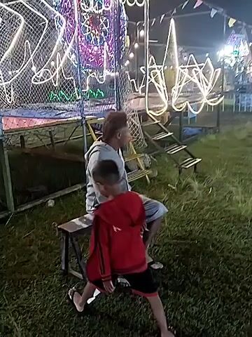 The video shows a night scene at an outdoor fair or carnival. There are people walking around and colorful lights illuminating the area. One shot shows a man sitting on a bench with a child standing near him. In the background are light displays, rides, and game booths, strung with colorful pennant banners. People are visible at a game booth, interacting with the vendor. The atmosphere appears lively and festive.