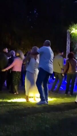 In the video, a group of people are dancing outdoors at night. The foreground focuses on a man and a woman dancing together. The man is wearing a white shirt and blue jeans, while the woman is wearing a white dress. They are holding hands and appear to be engaged in a slow dance. Several other people are visible in the background, also dancing and socializing. The environment is dark, with trees and foliage suggesting a park or an outdoor venue. Blue and white lights are illuminating the area, possibly from stage lighting or decorative elements. The overall atmosphere appears to be festive and relaxed.