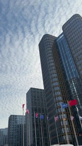 The video shows a low-angle view of several tall buildings against a partly cloudy sky. The clouds are small and white, creating a textured appearance. Various flags representing different nations are visible at the base of the buildings. The buildings are modern with glass and metal exteriors. The video appears to be taken outdoors on a bright day.