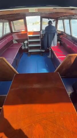 The camera is located inside of a wooden boat that is traveling on a body of water. There is a person at the wheel of the boat, and red benches are on either side. The boat's wake is visible in the water. The Italian flag is on the back of the boat. There are buildings and trees on land in the distance.
