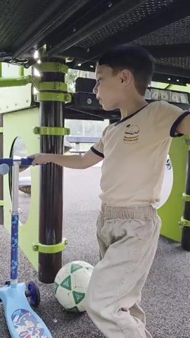 A boy stands near a playground structure and a scooter. He is wearing a tan t-shirt and khaki pants. A soccer ball is on the ground next to him. The boy is looking down at the ball and then up at the camera.