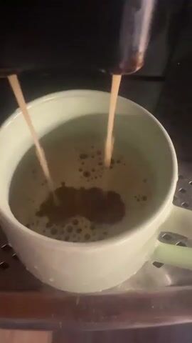 This is a close-up, stationary shot of an espresso machine dispensing two streams of dark liquid into a light green mug. The liquid is creating a frothy, bubbly surface in the mug. The espresso machine's spouts are visible at the top of the frame, and the mug is resting on a metal grate. The lighting is dim, creating a warm glow.