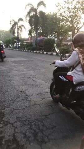 A person wearing a helmet and a brown hijab rides a black motorcycle on a paved road. The motorcycle has a license plate. The background shows trees and a green truck. The sun is setting or rising, casting a warm light.