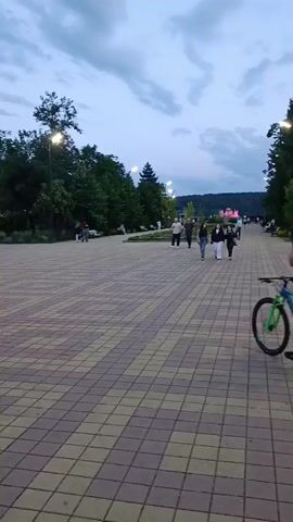 A person rides a bicycle down a paved walkway in a park. In the background, several other people are walking. A large sign with the word "MAIKO" in illuminated white letters is visible on the right side of the frame. Trees and streetlights line the walkway. The sky is cloudy and appears to be twilight.