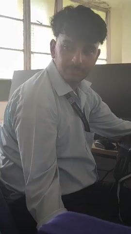 A man with dark hair and a mustache is sitting in front of a computer. He is wearing a light blue collared shirt with a lanyard around his neck. The lanyard has text on it that reads "College Dharamshala". The man is looking off to the side and slightly upwards. In the background, there is a window with a grid-like pattern and a dark computer monitor. The lighting in the room appears to be coming from the window.