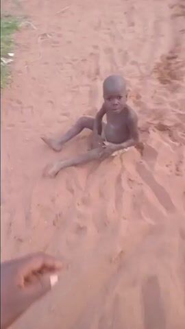 Here is a description of the video:

The video shows a small child sitting on a sandy surface, looking at the camera, and turning his head to look behind him. The person holding the camera moves around the child, walking around him and looking at him from different angles. The child does not move. At one point, a white cloth or paper is visible on the sand near the child. The sand appears reddish brown. The background includes some grass and trees. The child's skin appears dark.